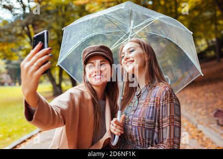 Outdoor-Porträt von zwei jungen Frauen, die Selfie mit Telefon unter Regenschirm während regen im Herbst Park. Stockfoto