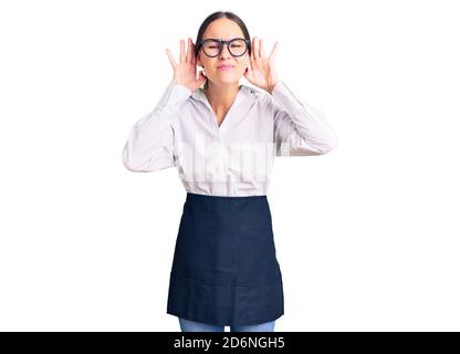 Schöne Brünette junge Frau trägt professionelle Kellnerin Schürze versuchen, beide Hände auf Ohr Geste hören, neugierig auf Klatsch. Hörproblem, taub Stockfoto