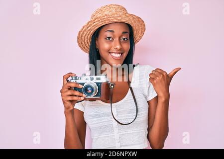 Junge afroamerikanische Frau trägt Sommerhut halten Vintage-Kamera zeigt auf den Rücken hinter mit Hand und Daumen nach oben, lächelnd zuversichtlich Stockfoto