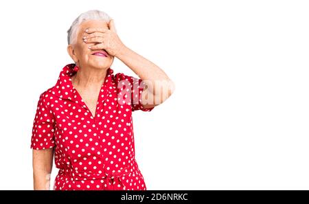 Ältere schöne Frau mit blauen Augen und grauen Haaren trägt ein rotes Sommerkleid lächelnd und lachend mit Hand auf Gesicht bedeckende Augen für Überraschung. blin Stockfoto
