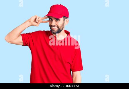 Junge gutaussehende Mann mit Bart tragen Lieferung Uniform tun Frieden Symbol mit Fingern über Gesicht, lächelnd fröhlich zeigt Sieg Stockfoto