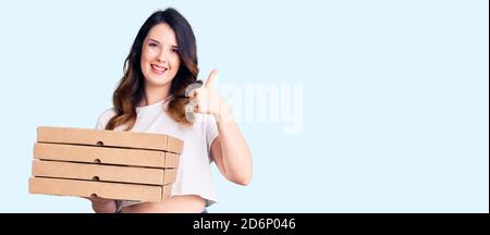 Schöne junge Brünette Frau hält Lieferung Pizza Box lächelnd glücklich und positiv, Daumen nach oben tun ausgezeichnet und Genehmigung Zeichen Stockfoto