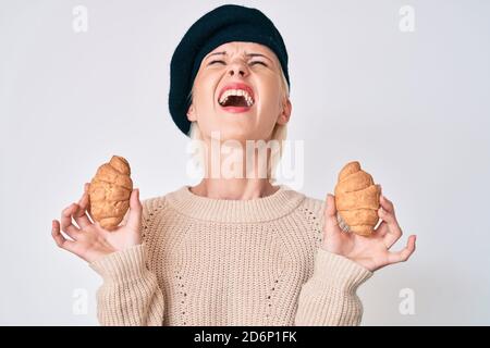 Junge kaukasische Frau im französischen Look mit Baskenmütze, die Croissants hält, wütend und wütend schreiend frustriert und wütend, schreiend mit Wut, die aufschaut Stockfoto