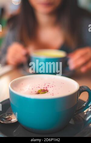 Nahaufnahme eines schäumenden Rose Latte Kaffees mit Blütenblättern In einem blauen Becher und Frau im Hintergrund Stockfoto