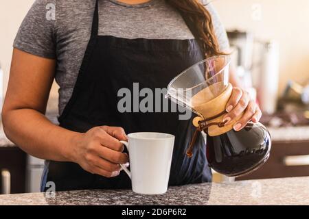 Hispanische Barista gießt Kaffee in eine weiße Tasse in der Familienküche. Schwarze Frau hält gießen über Kaffeekanne am Morgen Stockfoto