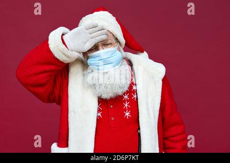 Kranker Weihnachtsmann trägt Gesichtsmaske mit Fieber oder Kopfschmerzen auf rotem Hintergrund. Stockfoto