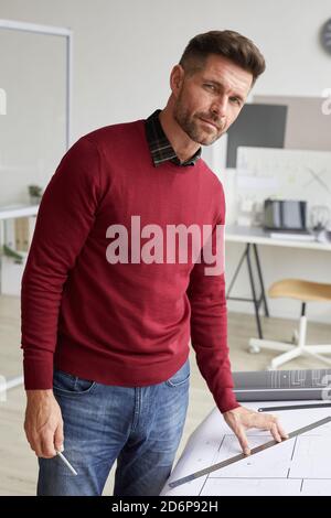 Vertikales Porträt von reifen bärtigen Architekten Blick auf Kamera während Am Schreibtisch am Arbeitsplatz stehen Stockfoto