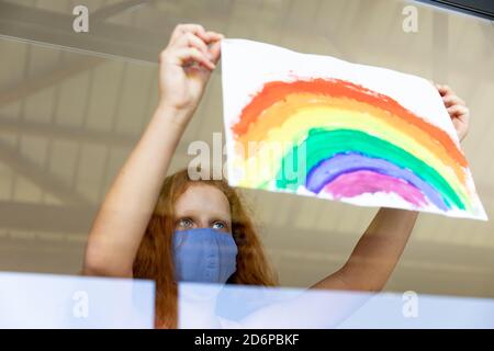 Mädchen tragen Gesichtsmaske kleben Regenbogen Malerei an das Fenster Stockfoto