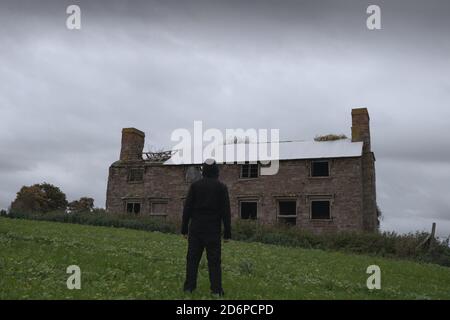 Ein gespenstisch verlassenes Haus, bedeckt von Vegetation. Mit einer Kapuzenfigur, die auf dem Land steht Stockfoto