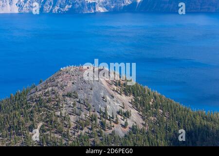 Der Gipfel der Wizard Island mit einem Pfad, der nach oben führt Zum Krater über Steinfelsen und an toten Bäumen vorbei Mit dem pulsierenden blauen Wasser des Crater Lake in der Stockfoto