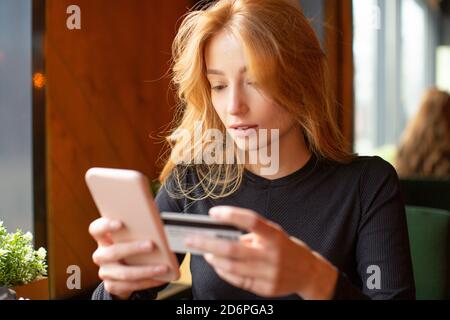 Rothaarige junge Frau, die Kartenzahlung über das Handy macht, um Rechnungen in einem Café zu bezahlen. Nahaufnahme. Stockfoto