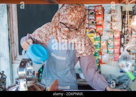 Das Mädchen im Hijab in der Schürze gießt Wasser aus Der Wasserkocher, um Getränke für den Kunden am Wagen zu machen Stall Stockfoto