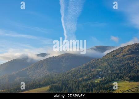 Niedrige bewaldete Berge. Sonniger Morgen und leichter Nebel. Eine ungewöhnliche Wolke und mehrere Häuser auf der Piste Stockfoto