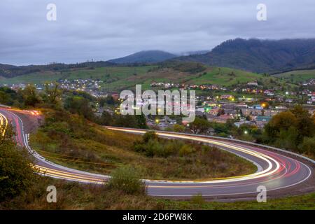 Wolkiger Abend auf einer Bergstraße. Scheinwerferspuren von schnell fahrenden Autos. Ein kleines Dorf erstrahlt mit Lichtern im Tal Stockfoto