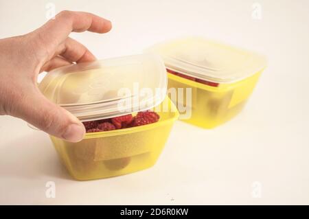 Eine Frauenhand schließt die Lunchbox, das Foto zeigt eine Hand und zwei gelbe Lunchboxen mit Himbeeren Stockfoto