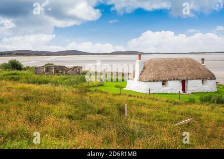 Traditionell gebaute weiße Hütte mit Strohdach, neben der türkisfarbenen Bucht, mit stürmischen bewölkten dunklen Himmel über.Insel North Uist, Schottland Stockfoto