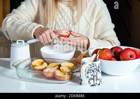 Weibliche Hände schneiden einen Apfel und bereiten ihn zum Backen, auf dem Tisch in einer weißen Schüssel mit Äpfeln, hausgemachtes Backkonzept für den Urlaub Stockfoto