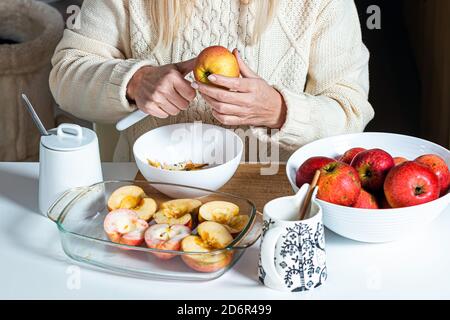 Weibliche Hände schneiden einen Apfel und bereiten ihn zum Backen, auf dem Tisch in einer weißen Schüssel mit Äpfeln, hausgemachtes Backkonzept für den Urlaub Stockfoto