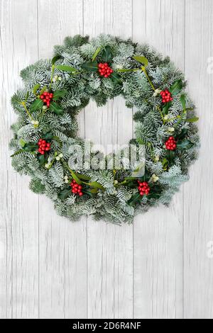 Natürliches Wintergrün Kranz für Weihnachten mit schneebedeckter Fichte Tanne, Stechpalme & Mistel auf rustikalem weißem Holz Hintergrund. Traditionelle Komposition Stockfoto