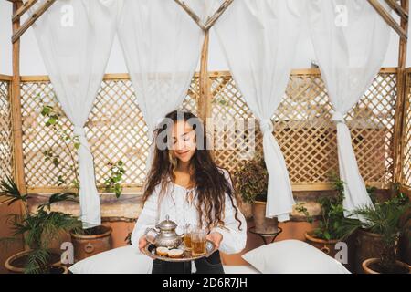 Schöne junge orientalische Frau hält Tablett mit traditionellen marokkanischen Minztee, Cookies und Silber Teekanne. Gastfreundschaft in Marokko. Stockfoto