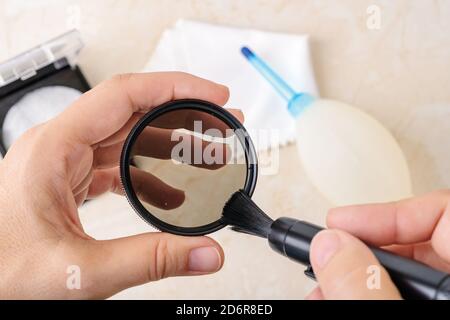 Hände, die den Staub von einem Kamera-Polarisationsfilter mit der Linsenstiftbürste abfegen. Entfernung von Staub und Fingerabdrücken von Fotogeräten. Stockfoto