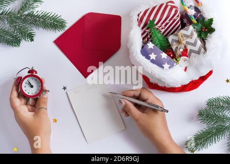 Weihnachtskarte mit Frauenhänden Schreiben Brief an den Weihnachtsmann, halten Wecker und roten Hut gefüllt verschiedene Geschenke auf einem hellgrauen Hintergrund, Cop Stockfoto