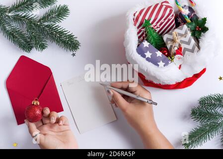 Weihnachtskarte mit Frauenhänden Schreiben Brief an den Weihnachtsmann, halten Wecker und roten Hut gefüllt verschiedene Geschenke auf einem hellgrauen Hintergrund, Cop Stockfoto