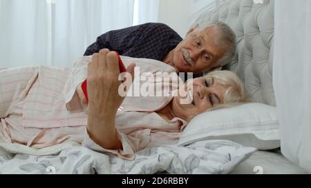 Ältere ältere Pärchen tragen Schlafanzug auf dem Bett liegend auf Handy lachen und Spaß haben. Ältere kaukasische Großeltern, Ehemann und Ehefrau nach dem Aufwachen machen Online-Shopping Stockfoto