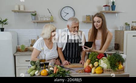 Ältere Großeltern Paar mit Enkelin in der Küche. Ältere Mann und Frau hören Rezept, Ratschläge von Mädchen mit digitalen Tablette gesunde Ernährung. Rohkost Essen Diät. Welt vegan Tag Stockfoto