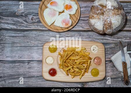 Draufsicht auf einen Tisch mit Pommes Frites mit verschiedenen Saucen, einen Tisch mit Spiegeleiern in Herzform und ein rustikales Dorfbrot Stockfoto