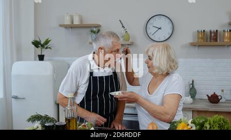 Ältere Großeltern in moderner Kücheneinrichtung. Ältere Frau Fütterung Mann mit rohen Sprossen Buchweizen mit Nüssen. Öko-Lebensmittel essen Ernährung. Gesunder reifer Familienleben. Welt vegan Tag Stockfoto