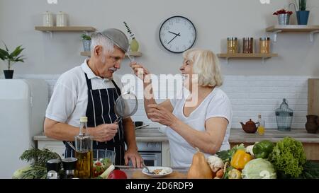 Glückliches Seniorenpaar tanzen beim Kochen zusammen in der Küche mit frischem Gemüse und Obst zu Hause. Ältere Großmutter und Großvater machen einen lustigen Tanz mit Sportschuhen. Gesunde, reife Familie Stockfoto