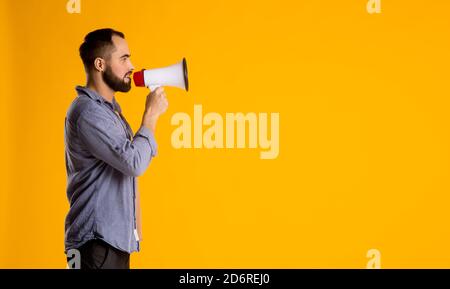 Ein bärtiger Mann in einem lässigen Hemd mit Lautsprecher In seinen Händen schreit in ein Megaphon auf einem gelben Hintergrund Stockfoto