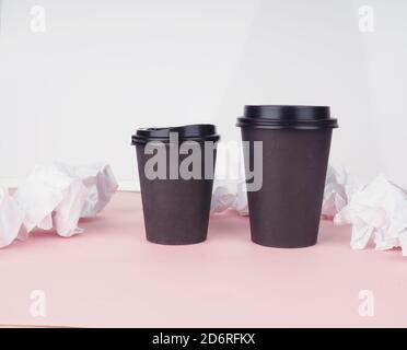 Zwei braune Pappkaffeetassen auf einem rosa Hintergrund, daneben zerknittertes Papier. Stockfoto