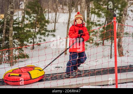 Junge mit Schlauch steigt auf einem Travelator auf den Berg. Kind hat Spaß an der Schneeröhre. Junge reitet einen Schlauch. Winterspaß für Kinder Stockfoto