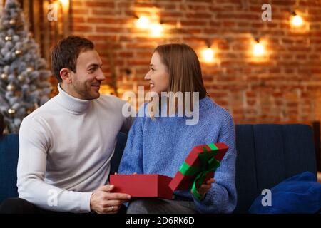 Schöner Mann, der seiner schönen Frau ein Geschenk übergibt und lächelt. Schöne junge Paar zu Hause genießen Zeit zusammen. Winterurlaub Stockfoto