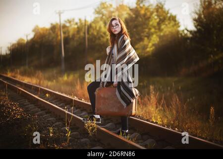 Eine schöne junge Frau in einem gestreiften Schal reist mit einem Retro-Koffer in ihren Händen und überquert die Eisenbahn, beleuchtet von den Strahlen des Sonnenlichts. Li Stockfoto