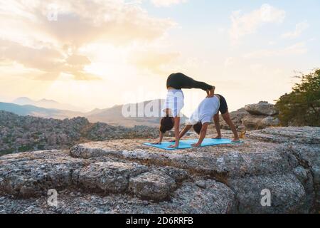 Ganzer Körper eines nicht erkennbaren Paares, das Akro-Yoga praktiziert, und ein Mann, der nach unten schauende Hundepose macht, während eine Frau in Moun einen Handstand auf dem Felsen aufführt Stockfoto