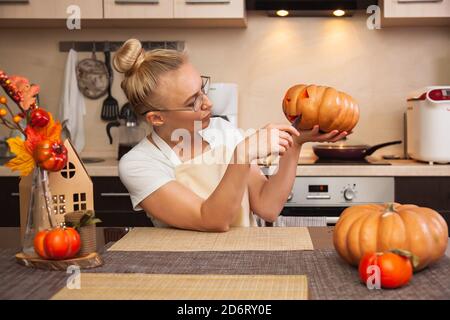 Frau in der Küche schnitzt einen Kürbis für Halloween in einem Raum mit Herbstdekor und einem Lampenhaus. Gemütliches Zuhause und Vorbereitung auf Halloween. Stockfoto