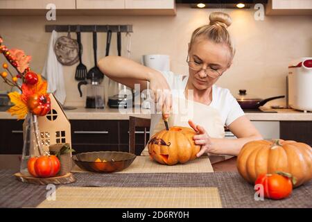 Frau in der Küche schnitzt einen Kürbis für Halloween in einem Raum mit Herbstdekor und einem Lampenhaus. Gemütliches Zuhause und Vorbereitung auf Halloween. Stockfoto