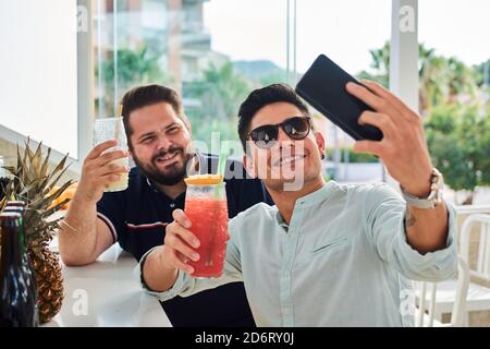 Glückliche junge Männer in lässigen Outfits lächeln und Selfie machen Auf dem Smartphone, während Sie frische Smoothies im tropischen Café trinken Stockfoto