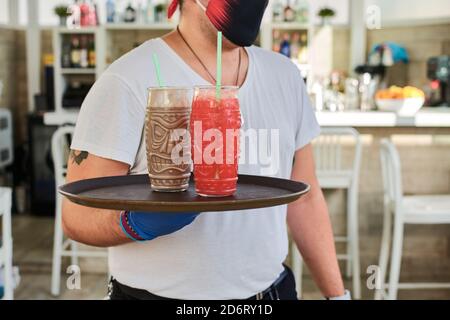 Crop anonymen jungen Kellner in T-Shirt und Maske tragen Tablett mit frischen Getränken Gläser im modernen Café Stockfoto