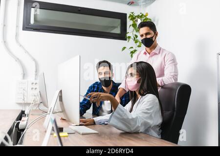Anonymer Unternehmer in Maske zeigt Projekt auf Desktop-Computer während Interaktion mit jungen Kollegen im Büro Stockfoto