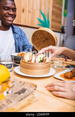 Crop lächelnde multiethnische Freunde versammeln sich am Tisch im Café Und Abendessen mit leckeren Sandwiches Stockfoto