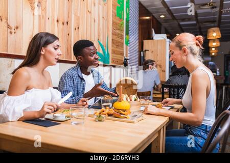 Positive junge multirassische Freunde essen leckere Gerichte und Burger während Treffen im modernen Restaurant Stockfoto