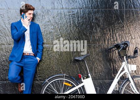 Serious männlich Executive in formellen Anzug lehnte an der Wand, während Internet auf Handy sprechen und mit der Hand in der Tasche stehen In der Nähe von Elektrofahrrädern Stockfoto