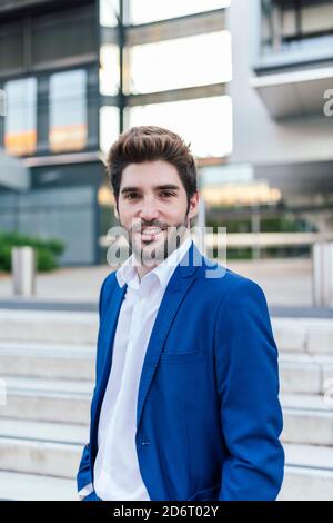 Junger lächelnder bärtiger Geschäftsmann in eleganter Kleidung mit modernem Haarschnitt Blick auf die Kamera in der Nähe von Gebäude mit Treppe in der Stadt Stockfoto