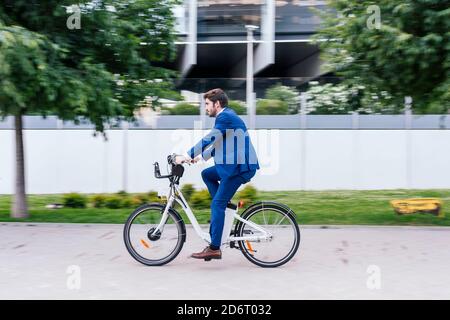 Junger lächelnder Mann in elegantem Anzug, der mit dem Elektrofahrrad fährt Bürgersteig beim Blick weg Stockfoto