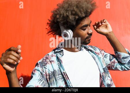 Junger männlicher bärtiger afroamerikanischer Mann mit Afro-Frisur und Geschlossene Augen hören Musik in Kopfhörern in der Nähe der roten Wand In der Stadt Stockfoto