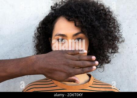 Anonymer afroamerikanischer Freund, der das Gesicht der jungen aufmerksam bedeckt Weibchen mit Afro Frisur in der Nähe Zementwand beim Betrachen Kamera Stockfoto